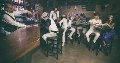 Bartender serve drinque no balcão enquanto a banda ocupa as mesas altas