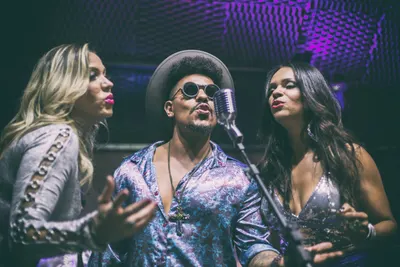 Três vocalistas em volta do microfone sob luz roxa